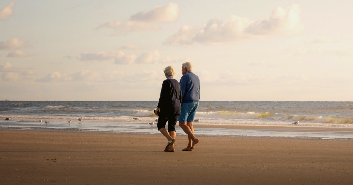 Un cuplu de seniori se plimbă liniștit pe plajă, la apus — imagine a unei pensionări demne, construite din timp.